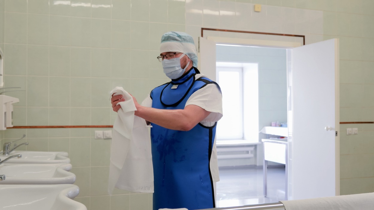 Masked medical worker wearing protective blue lead apron and cap stands in clean tiled hospital washroom while holding white coat near sinks with open doorway and soft clinical light